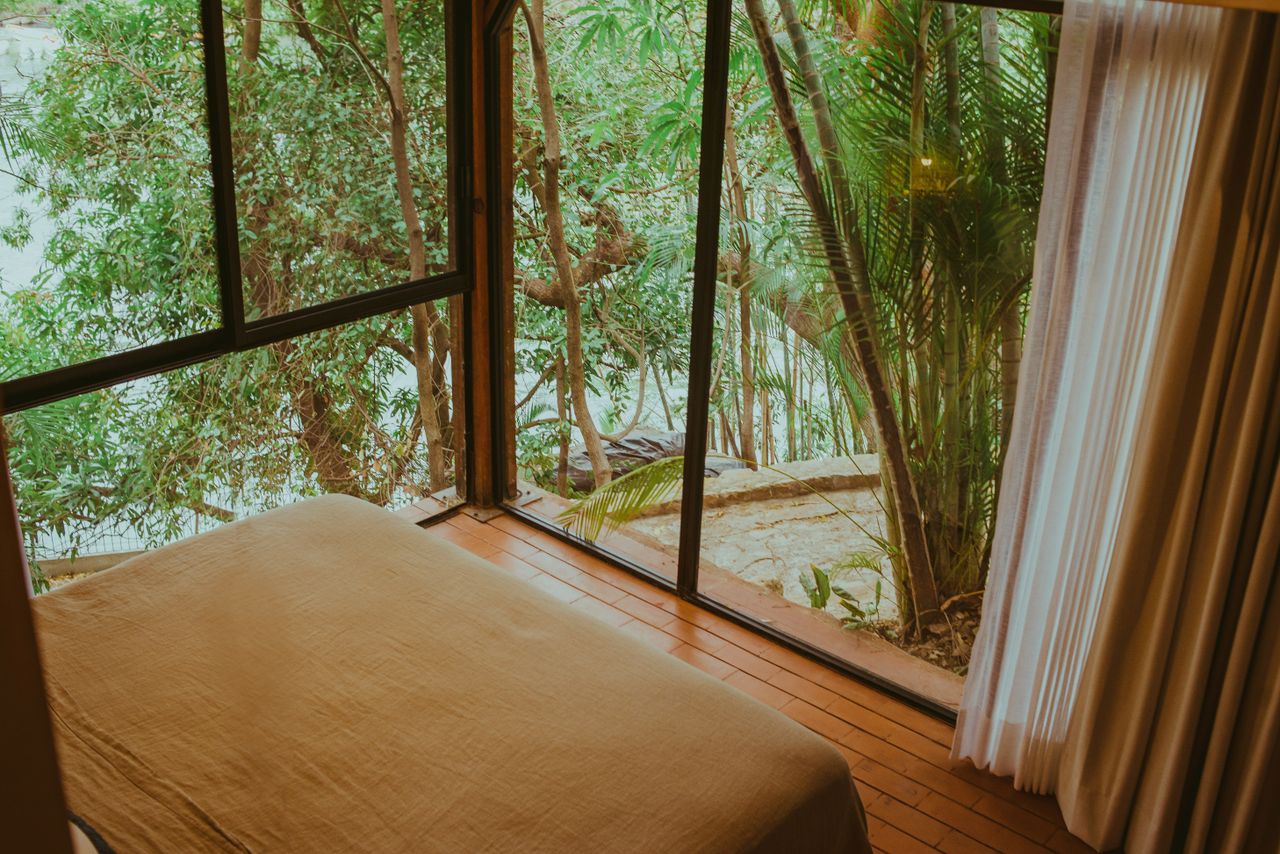A cozy bedroom overlooking lush tropical greenery through a large window, creating a relaxing ambiance.