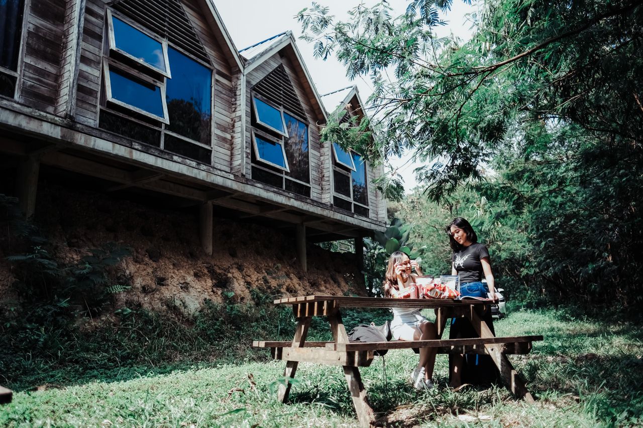 Two people enjoy a picnic near a rustic wooden cabin amidst lush greenery on a sunny day.